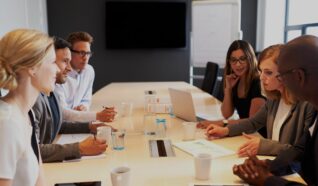 photo of a group of people sat around a desk listening to 1 person speak
