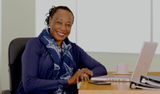 photo of a woman sat at a desk smiling at the camera with their laptop in front of them