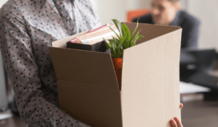 photo of a person stood in an office with a box of paperwork and a plant